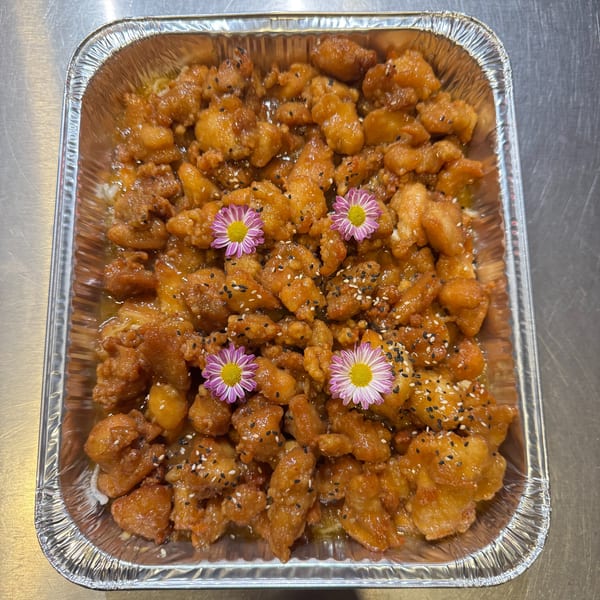 Sweet and sour chicken in a foil tray, garnished with sesame seeds and purple flowers.