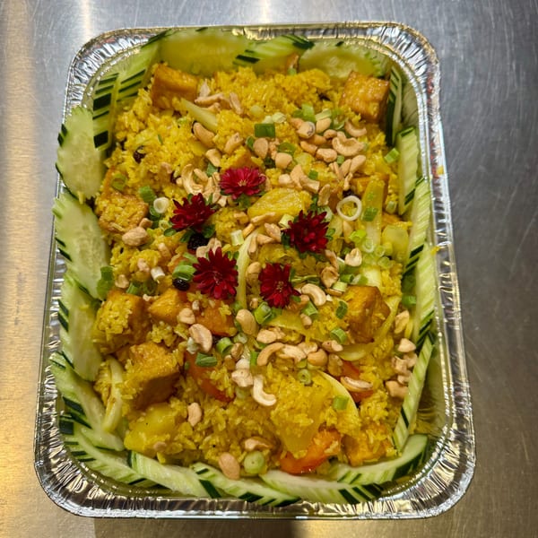 Yellow curry fried rice with tofu, cashews, vegetables, and red flowers in a foil container.