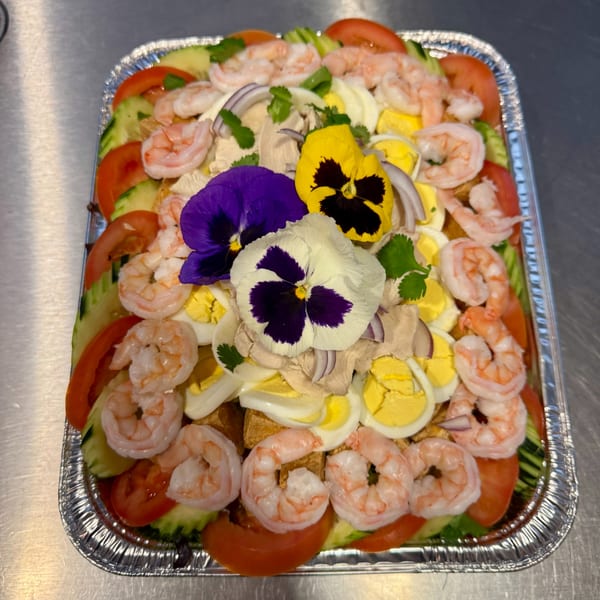 A vibrant salad with shrimp, tomatoes, cucumbers, eggs, and edible flowers in a foil tray.
