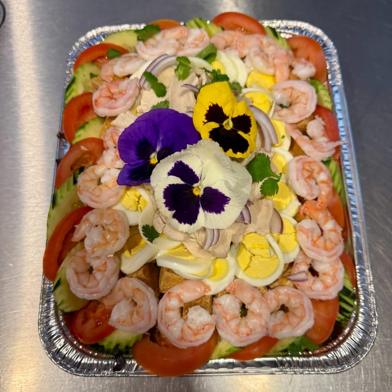 A vibrant salad with shrimp, tomatoes, cucumbers, eggs, and edible flowers in a foil tray.