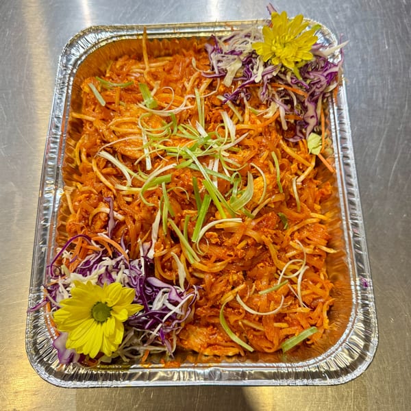 Spicy orange noodles with shredded cabbage, carrots, and green onions in a foil tray.